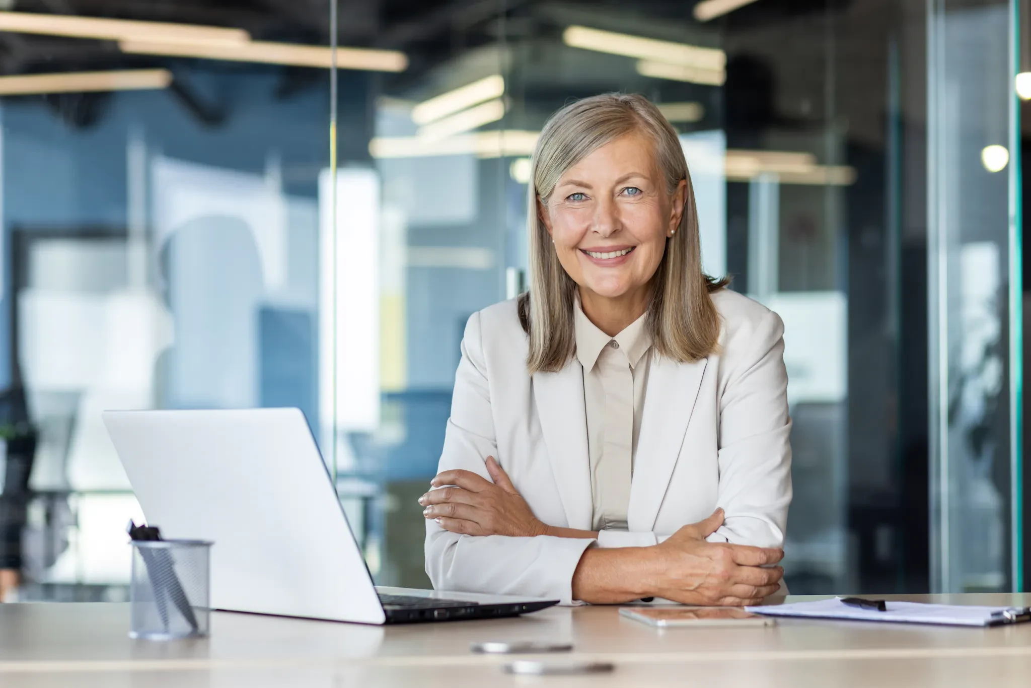 Confident businesswoman in a light suit smiling at her desk with a laptop in a modern office setting.