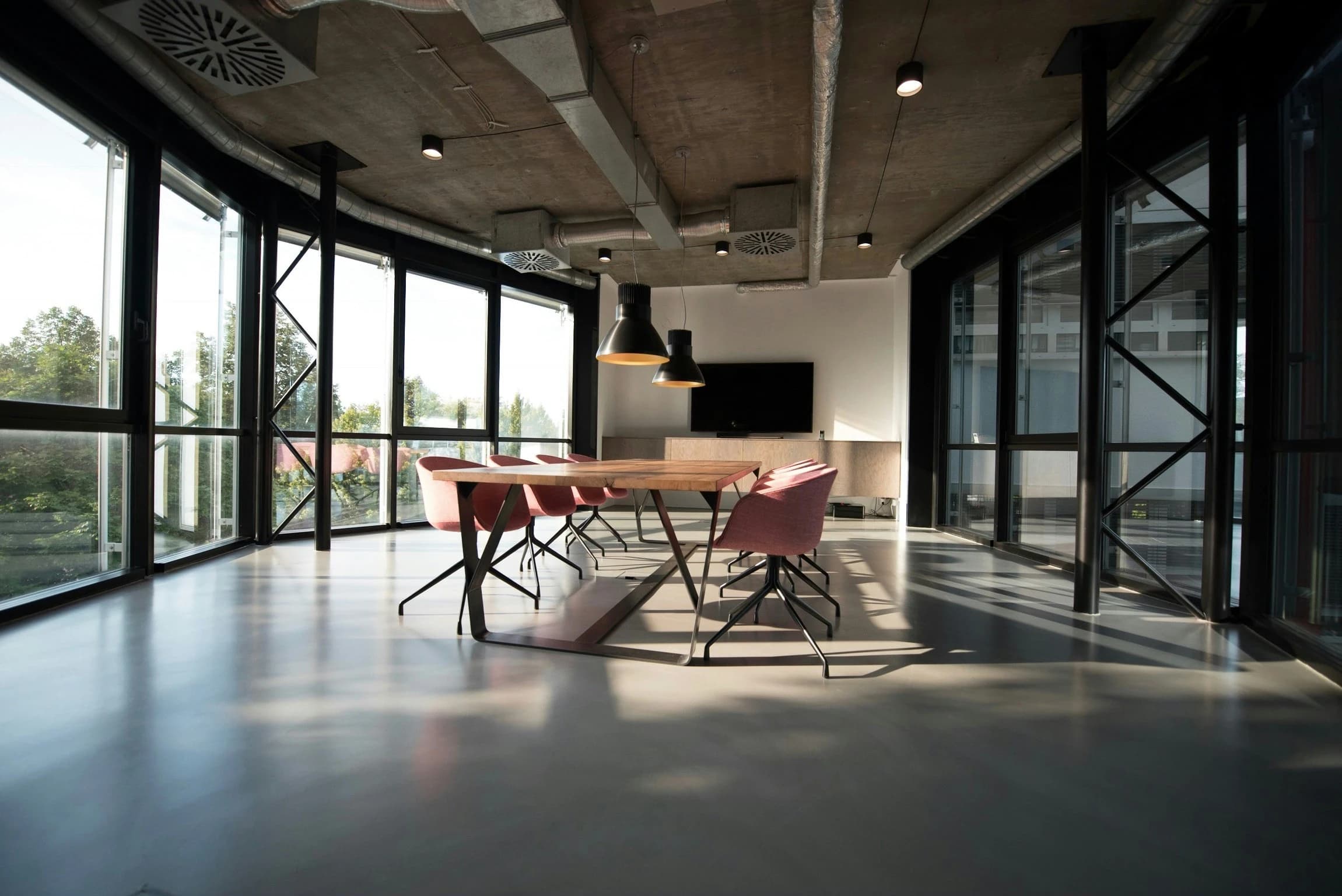 Modern conference room with large windows, a wooden table, pink chairs, and industrial lighting.