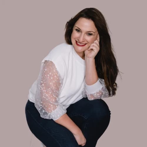 Joleen, smiling warmly, wearing a white blouse with lace sleeves and dark jeans, posed confidently.