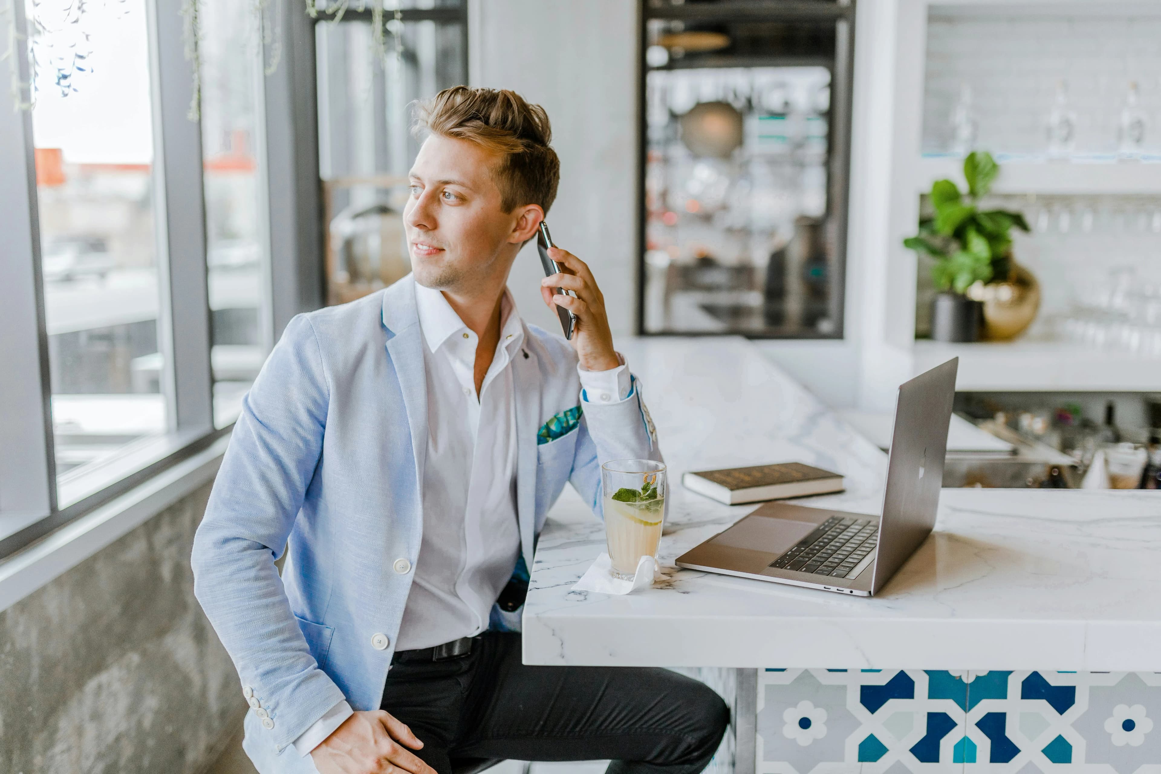Man in a light blazer sitting at a modern cafe with a laptop, drink, and phone, looking thoughtfully out the window