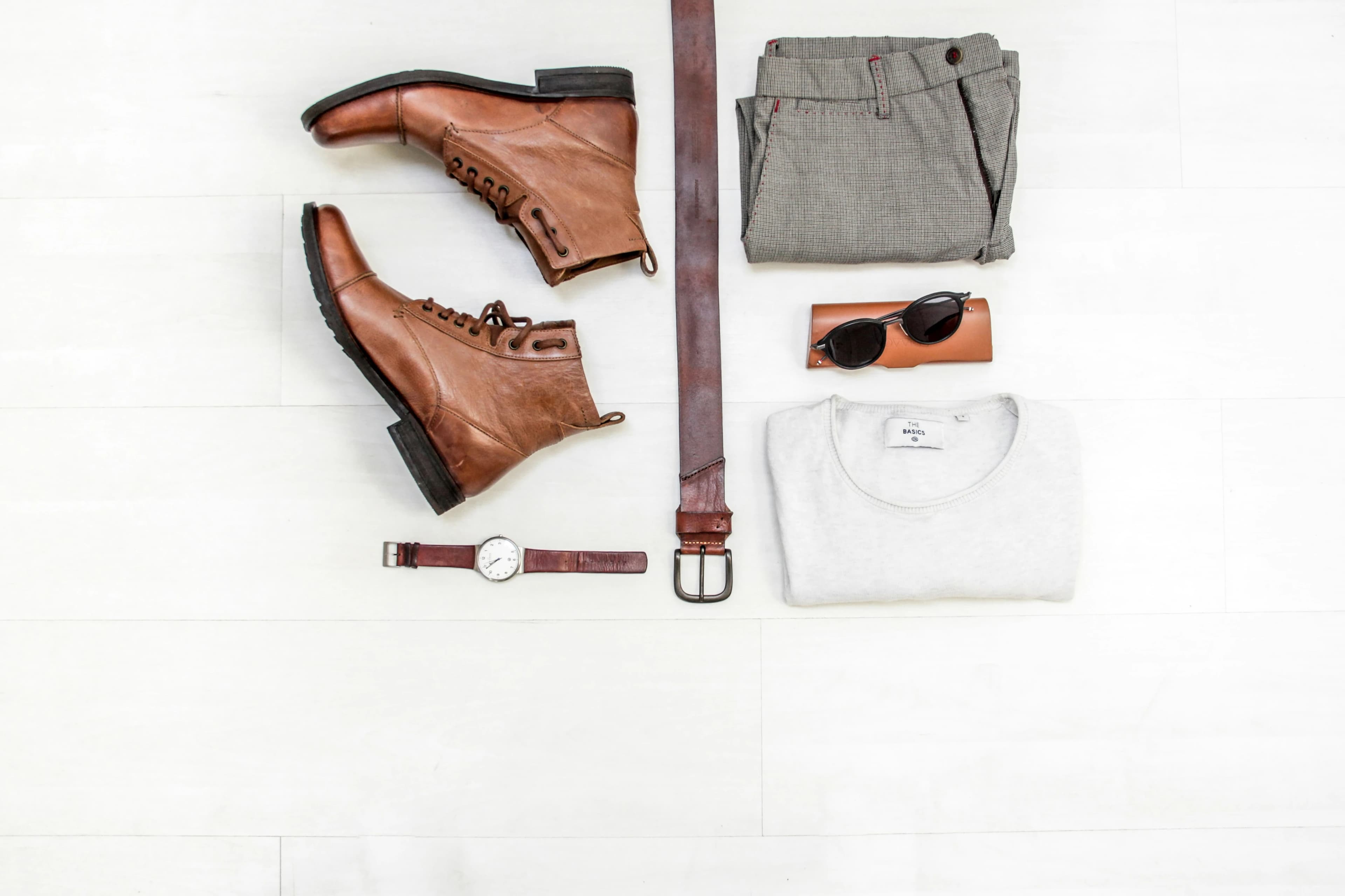 Flat lay of men's fashion items including brown leather boots, a belt, a gray folded shirt, trousers, sunglasses, and a wristwatch on a light background.